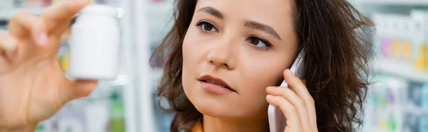 Brunette femme tenant bouteille avec des médicaments tout en parlant sur smartphone en pharmacie, bannière — Photo de stock