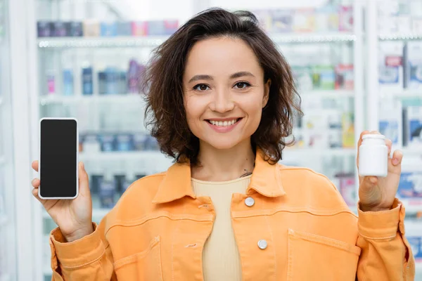 Femme gaie tenant bouteille avec des médicaments et smartphone avec écran blanc dans la pharmacie — Photo de stock