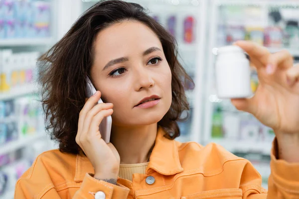 Брюнетка жінка тримає пляшку з ліками під час розмови на смартфоні в аптеці — стокове фото