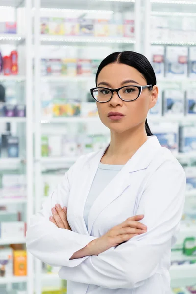 Asiático farmacêutico em óculos de pé com os braços cruzados na farmácia — Fotografia de Stock