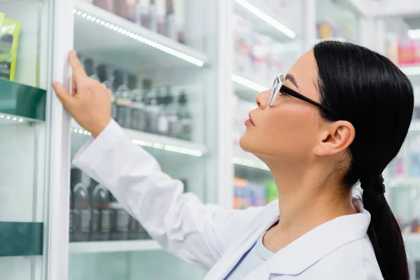 Vista laterale del farmacista asiatico in occhiali guardando farmaci su scaffali sfocati — Foto stock