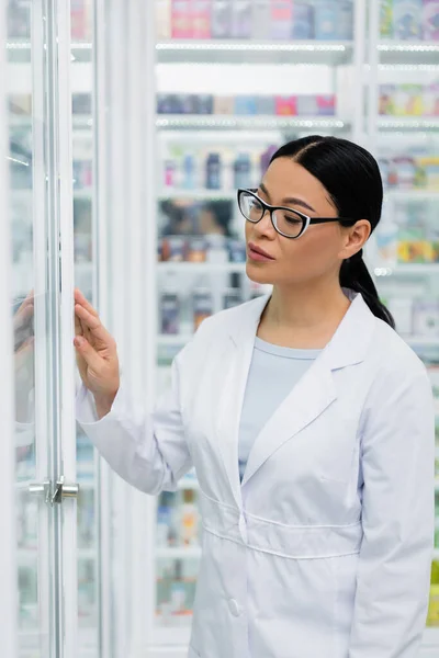 Farmacéutico asiático en gafas mirando la medicación en estantes borrosos - foto de stock