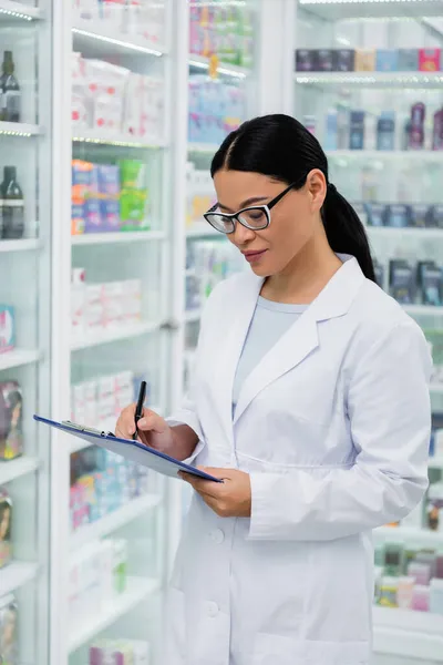 Farmacéutico asiático en gafas escritura en portapapeles en farmacia - foto de stock