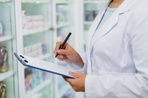 Cropped view of pharmacist in white coat writing on clipboard in drugstore — Stock Photo