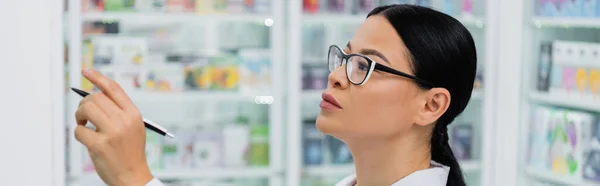Азіатський фармацевт в окулярах тримає ручку, вказуючи на наркотики, банер — стокове фото