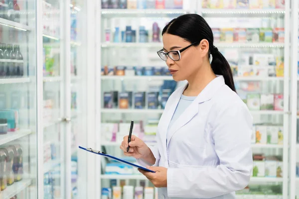 Asian pharmacist in white coat writing on clipboard in drugstore — Stock Photo