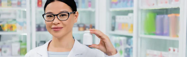 Farmacéutico asiático en gafas sonriendo mientras sostiene la botella con la medicación, bandera — Stock Photo