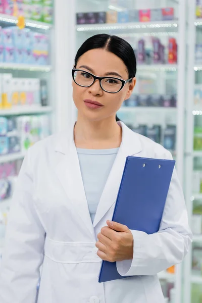 Asiático farmacêutico em óculos e enquanto casaco segurando prancheta — Fotografia de Stock