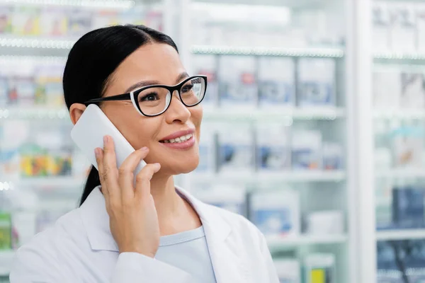 Cheerful asian pharmacist in glasses talking on smartphone — Stock Photo