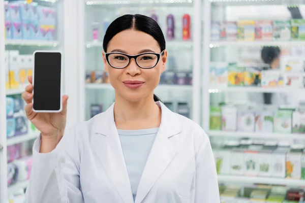 Asiatischer Apotheker mit Brille lächelt, während er sein Handy mit leerem Bildschirm in der Apotheke hält — Stockfoto