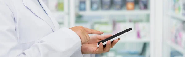 Cropped view of pharmacist in white coat pointing at smartphone, banner — Stock Photo