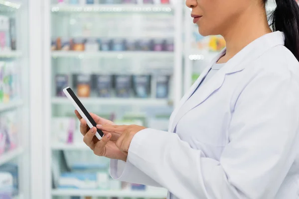 Cropped view of pharmacist in white coat pointing at cellphone in drugstore — Stock Photo