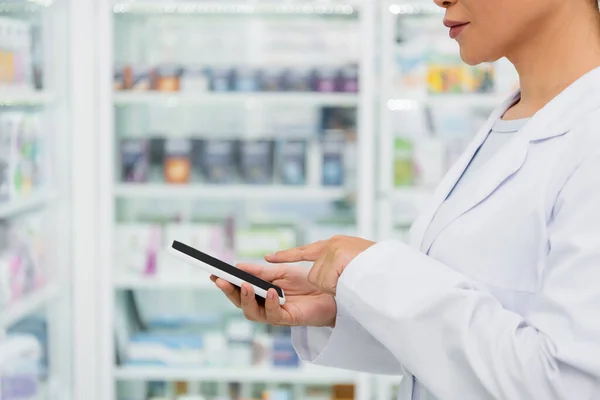 Vista cortada do farmacêutico em casaco branco apontando para o telefone celular na farmácia — Fotografia de Stock