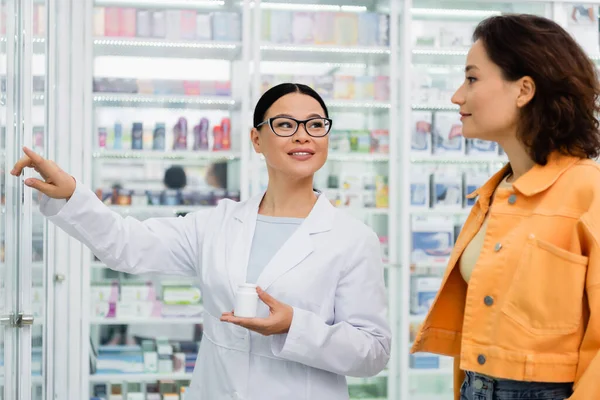 Cheerful asian pharmacist in glasses holding bottle with drugs and pointing away near brunette customer in drugstore — Stock Photo
