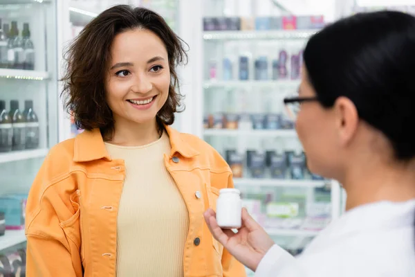 Farmacista offuscata in bicchieri che tengono bottiglia con farmaci vicino al cliente allegro in farmacia — Foto stock