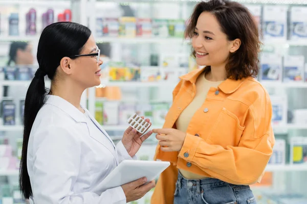 Asiatischer Apotheker in Brille mit Blisterverpackung und digitalem Tablet in der Nähe zufriedener Kunden in der Drogerie — Stockfoto