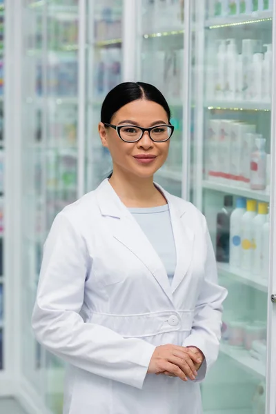 Веселый азиатский фармацевт в очках, стоящий с сжатыми руками в аптеке — стоковое фото