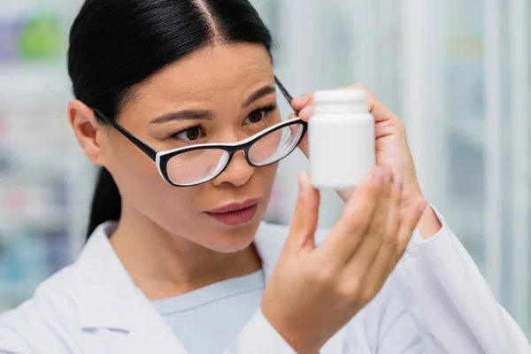 Brunette asian pharmacist adjusting glasses and looking at bottle with medication — Stock Photo