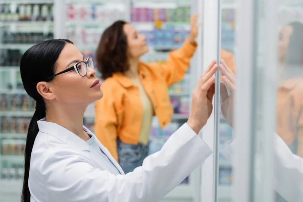 Asiático farmacêutico em óculos olhando prateleiras atrás de vidro perto de cliente borrado na farmácia — Fotografia de Stock
