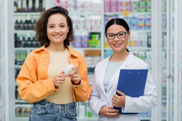 Asiático farmacéutico en gafas celebración portapapeles cerca alegre cliente con botella en droguería - foto de stock