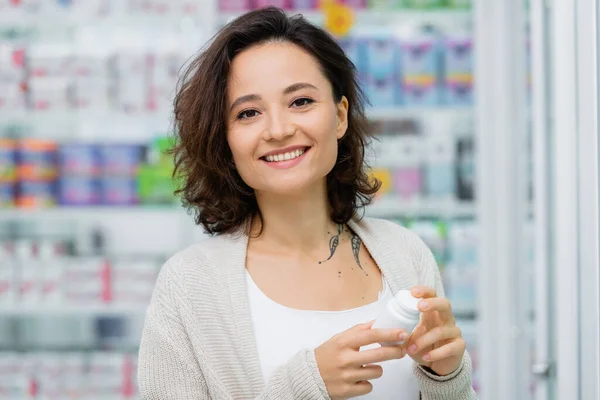 Fröhliche und tätowierte Frau mit Flasche mit Medikamenten in Drogerie — Stockfoto