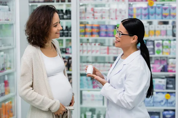 Азиатский фармацевт в очках держит бутылку с витаминами рядом с веселой беременной женщиной в аптеке — стоковое фото