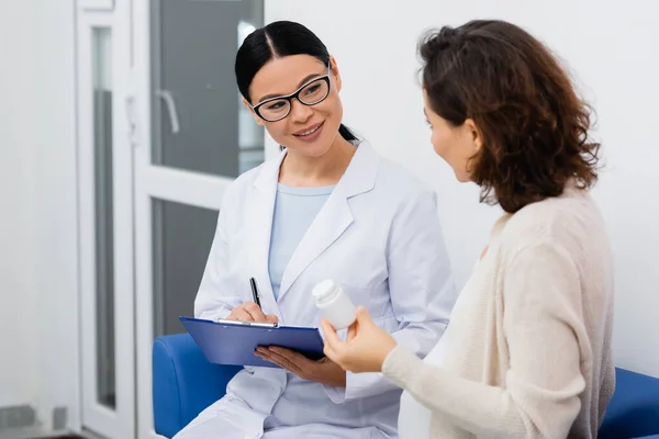 Cheerful asian pharmacist in glasses writing on clipboard near pregnant woman with bottle in drugstore — Stock Photo