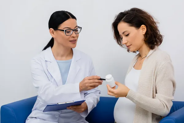 Asian pharmacist in glasses holding clipboard and pointing with pen at bottle near pregnant woman — Stock Photo