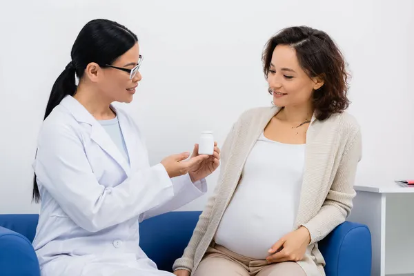 Asian pharmacist in glasses holding bottle with vitamins near cheerful pregnant woman sitting on sofa in drugstore — Stock Photo