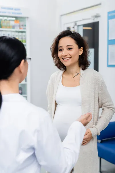 Femme enceinte avec tatouage souriant tout en regardant le pharmacien flou dans la pharmacie — Photo de stock