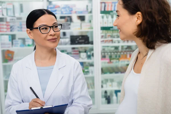 Asian pharmacist in glasses holding clipboard and pen near blurred and smiling pregnant woman — Stock Photo