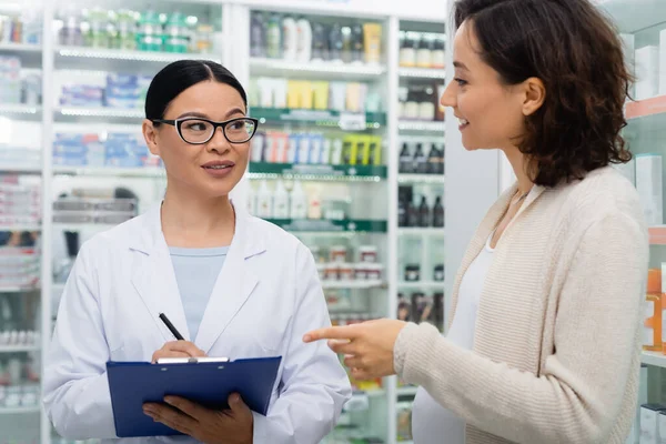 Asian pharmacist in glasses holding clipboard while talking with blurred and smiling pregnant woman — Stock Photo