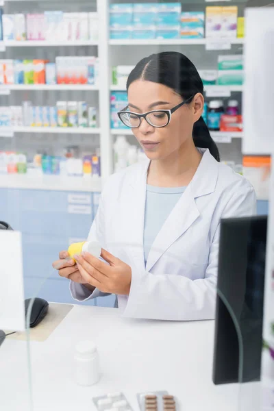 Brunette asian pharmacist in glasses and white coat looking at bottle with medication at counter — Stock Photo