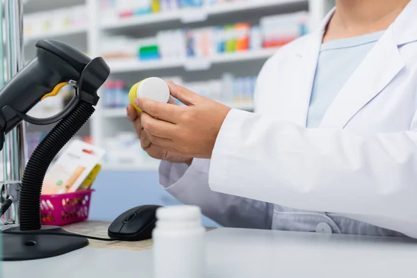 Vista ritagliata del farmacista in bottiglia bianca di scansione cappotto con farmaco al banco — Foto stock