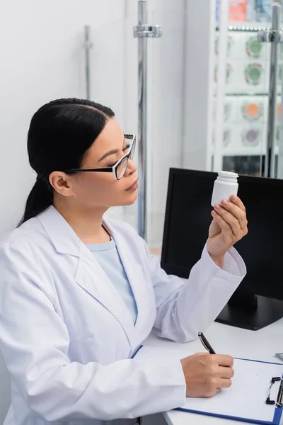 Asiático farmacêutico em óculos e casaco branco olhando para garrafa com pílulas enquanto escrevia na área de transferência — Fotografia de Stock