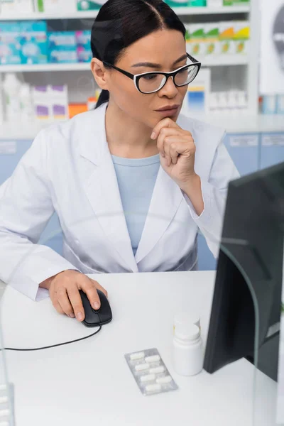 Brunette asian pharmacist in glasses and white coat using computer in drugstore — Stock Photo