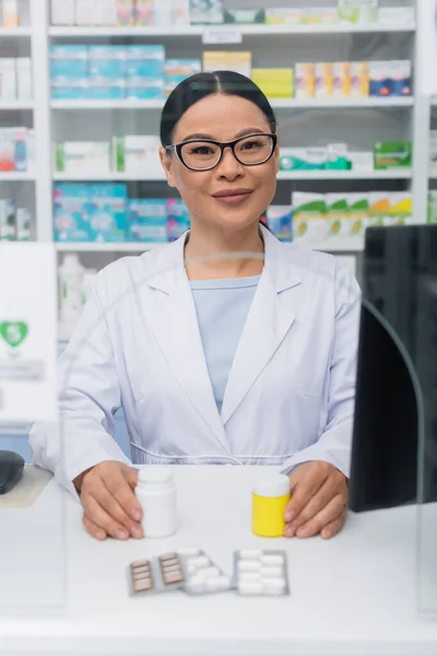 Alegre asiático farmacéutico celebración botellas con medicamentos cerca blister packs en escritorio en contador - foto de stock