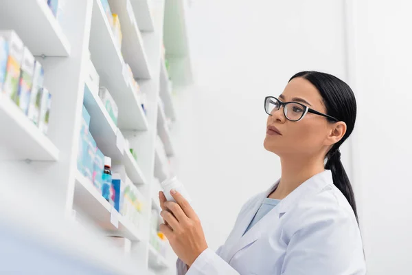 Vista de ángulo bajo de farmacéutico asiático en gafas mirando la medicación en farmacia - foto de stock