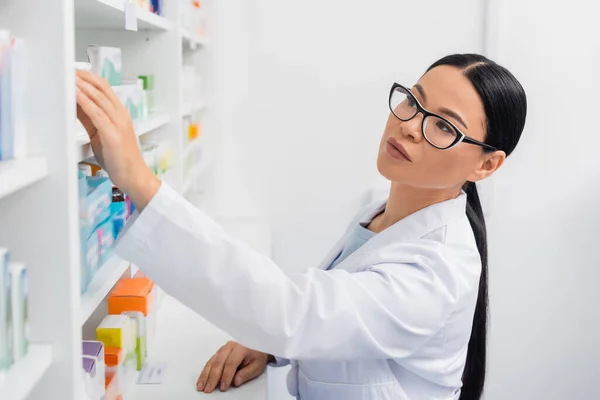 Farmacéutico asiático en gafas llegar a la medicación en farmacia - foto de stock