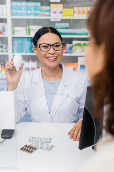 Alegre asiático farmacéutico celebración botella con medicación cerca borrosa cliente - foto de stock