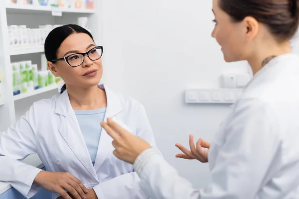 Desfocado farmacêutico no branco casaco gesto enquanto conversa com asiático colega no óculos — Fotografia de Stock