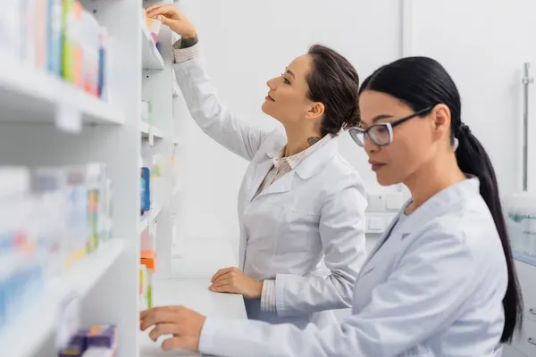 Cheerful pharmacist in white coat reaching carton box with medication near blurred asian colleague in glasses — Stock Photo