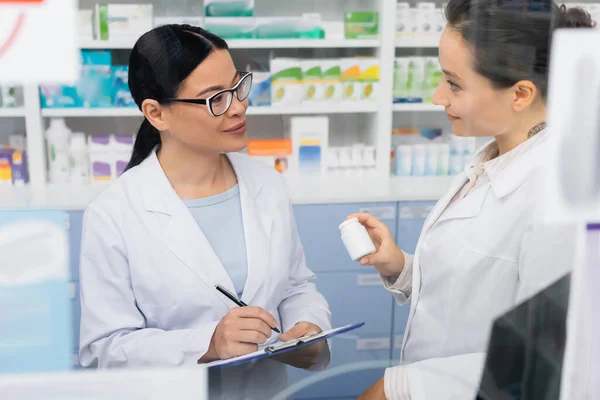 Tatuado farmacêutico no branco casaco segurando garrafa perto sorrindo asiático colega no óculos escrevendo no clipboard no drugstore — Fotografia de Stock