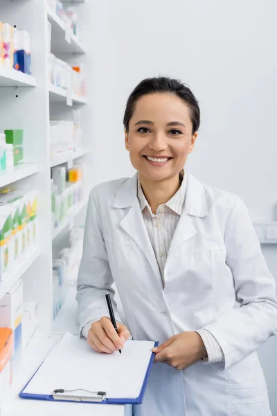 Farmacéutico sonriente con capa blanca escribiendo en el portapapeles - foto de stock