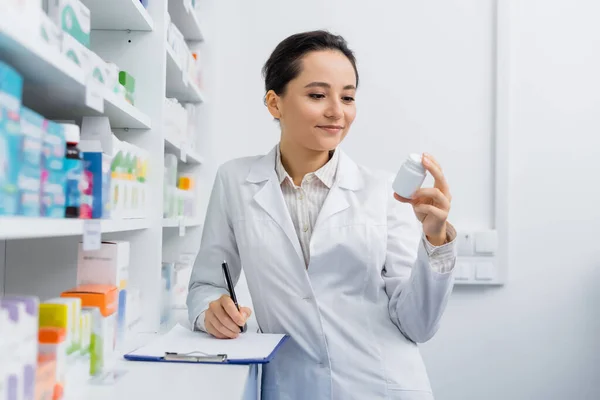 Pharmacist in white coat holding bottle with medication near clipboard — Stock Photo