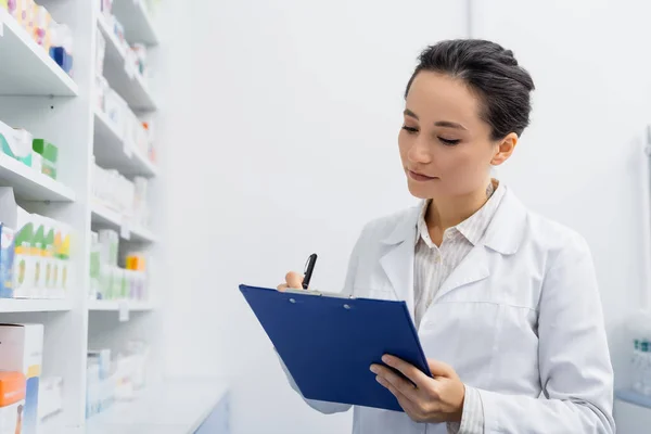 Tattooed pharmacist in white coat writing on clipboard in drugstore — Stock Photo