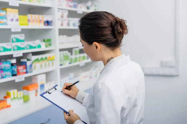 Tattooed apothecary in white coat writing on clipboard — Stock Photo