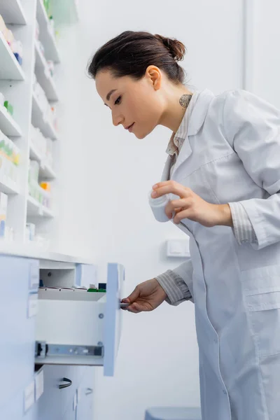 Boticario tatuado en abrigo blanco que sostiene la botella con la medicación mientras que abre el cajón en la farmacia - foto de stock