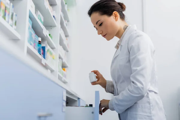 Pharmacien tatoué en manteau blanc tenant bouteille avec des médicaments lors de l'ouverture tiroir dans la pharmacie — Photo de stock