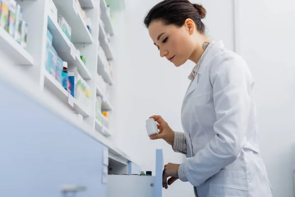 Tätowierter Apotheker im weißen Kittel hält Flasche mit Medikamenten in der Hand, während er Schublade in Drogerie öffnet — Stockfoto